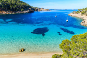 Aerial view of a bay in Ibiza, Spain including clear blue sea, two boats and the surrounding coast with trees