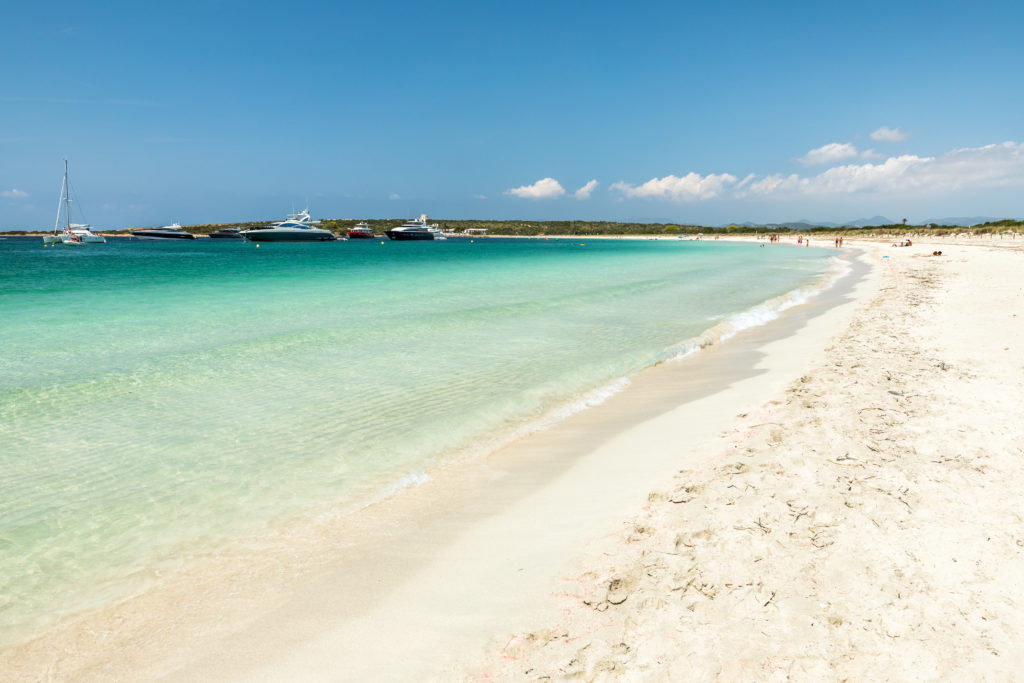 View of a beach on the island of s'Espalmador with white sand, clear seawater and a few boats on the background