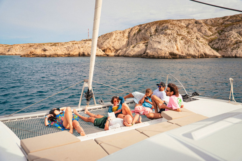 Group of people relaxing on board a catamaran anchored near a rocky coastline