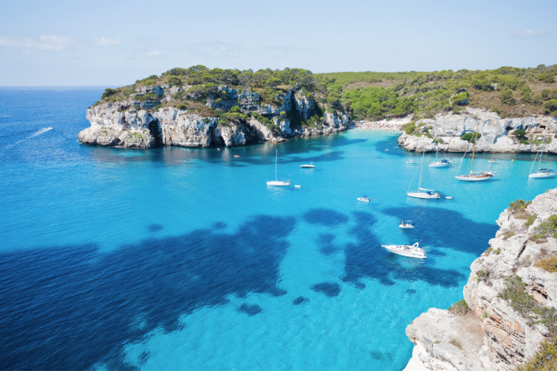 Boats and sailing yachts anchored in a turquoise cove surrounded by cliffs